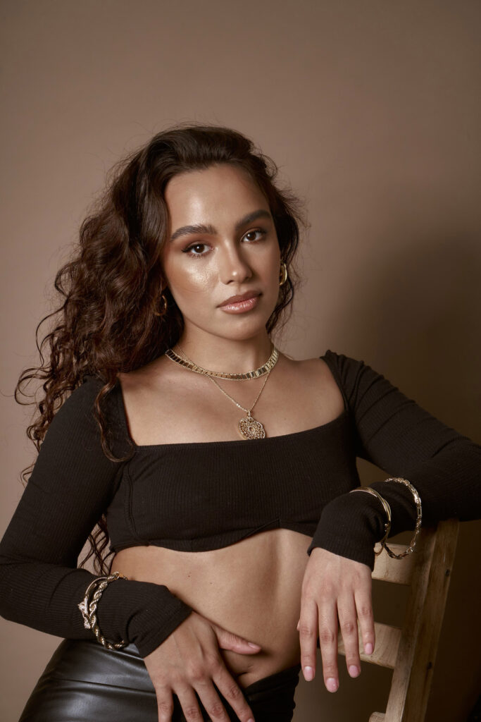 Abril Escalante a model with curly brown hair is posed against a muted brown backdrop. She wears a black off-shoulder top a gold necklace with a pendant and matching gold bracelets. Her makeup is subtle with emphasis on her eyes and lips. She rests her hand on a wooden chair.